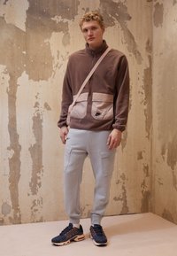 Hnedá fleecová mikina na zips s ružovými detailmi na vreckách, šedé cargo tepláky a námornícke tenisky s oranžovými akcentmi. Jednoduché pozadie.