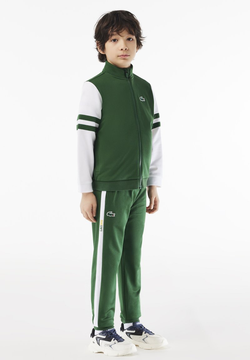 Zelena zip-up jakna s bijelim rukavima, s logotipom. Uske zelene hlače s bijelim bočnim trakama. Oba komada izrađena su od glatkog materijala, ležeran kroj.