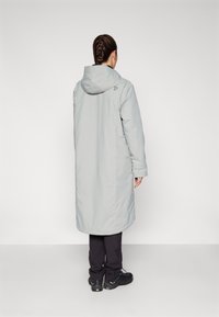 Parka imperméable grise avec une capuche, des manches longues et une coupe droite. Présente un logo à l'arrière près du cou et est associée à un pantalon foncé.