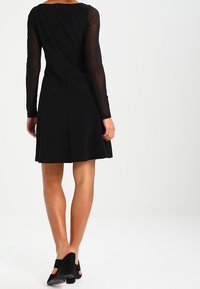 Frau, die ein schwarzes Kleid mit durchsichtigen langen Ärmeln und schwarzen Blockabsatzschuhen trägt, steht vor einem schlichten weißen Hintergrund.