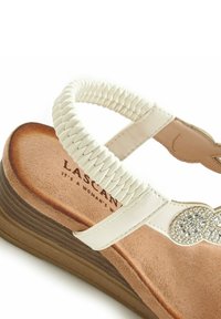 Witte sandaal met een gevlochten band en decoratieve strassaccenten, met een gestructureerd bruine platform en een zachte tan binnenzool.