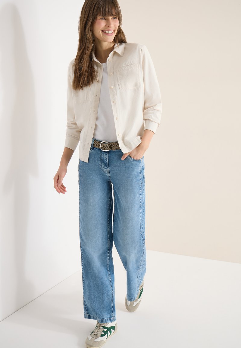 Femme souriante portant une chemise beige boutonnée, un haut blanc, un jean à jambes larges, une ceinture léopard et des baskets blanches marchant dans un espace neutre.