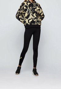Sudadera negra con patrón abstracto dorado y cordón, combinada con leggings negros ajustados que tienen acentos dorados y zapatillas deportivas negras.