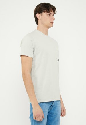 Giovane uomo di profilo che indossa una T-shirt grigio chiaro con taschino sul petto e jeans blu, su sfondo bianco.