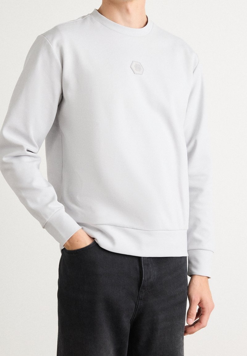 Homme portant un sweat-shirt gris clair avec un petit logo hexagonal sur la poitrine et un pantalon noir, debout avec une main dans la poche devant un fond uni.