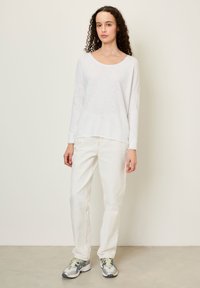 Kollase lokkis juustega naine seisab, kandes valget pika varrukaga särki, valgeid pükse ja halli jooksujalatsit, taustaks tavaline sein.