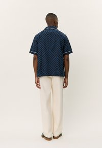 Camisa de manga curta em azul escuro com um padrão geométrico. Apresenta um colarinho, detalhes brancos e um corte descontraído, combinada com calças de cor creme.