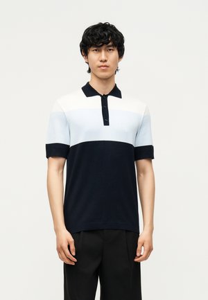 Giovane uomo con capelli corti e neri che indossa una polo a maniche corte con blocchi di colore nero, bianco e azzurro chiaro e pantaloni neri.