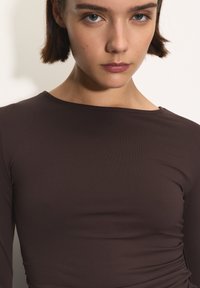 Brązowa, dopasowana bluzka z długim rękawem o prążkowanej fakturze. Okrągły dekolt z delikatnym marszczeniem z boku, co tworzy strukturalny wygląd.