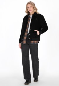 Kvinde iført en sort jakke over en ternet skjorte med sorte jeans og sorte støvler, der står med den ene hånd i jakke lommen.