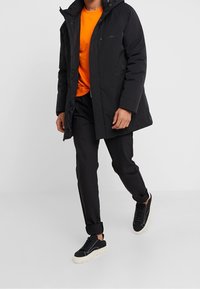 Sort polstret jakke med hætte, kontrasterende orange t-shirt, sorte bukser og sorte sneakers. Glat stof, funktionelt design, afslappet look.