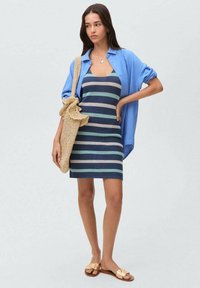 Abito a righe lavorato a maglia in blu, blu navy e crema; abbinato a una camicia azzurra, una borsa di paglia e sandali piatti dorati.
