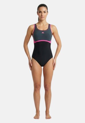 Donna in piedi rivolta in avanti che indossa un costume intero nero e grigio scuro con rifiniture rosa su uno sfondo bianco semplice.