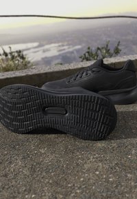 Pereche de pantofi sport negri pe trotuar, cu un pantof care arată modelul detaliat al talpii, fundal în aer liber neclar cu plante și vedere îndepărtată.