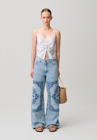 Giovane donna che indossa un top bianco con bordi in pizzo, jeans blu a gamba larga con ricamo blu scuro, tenendo una borsa beige.