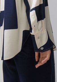 Long-sleeve shirt with cream and navy geometric pattern, featuring buttoned cuffs, smooth fabric, and a slightly curved hem.