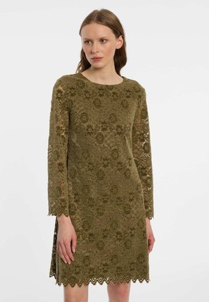 Frau trägt ein olivgrünes, langärmliges Spitzenkleid mit Blumenmuster, geschwungenem Saum und Ärmeln, steht vor einem einfarbigen hellen Hintergrund.