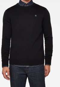 Schwarzer Pullover mit Rundhalsausschnitt, gerippten Bündchen und Saum. Verfügt über ein kleines Logo auf der Brust. Getragen über einem Jeanshemd.