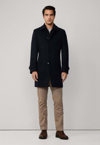 Uomo che indossa un cappotto nero con bottoni di lunghezza media, pantaloni beige e scarpe di pelle marroni, in piedi davanti a uno sfondo chiaro e semplice.