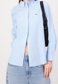 Ljusblå skjorta med vita ränder, tillverkad av bomull. Har en krage, långa ärmar och en liten logotyp på bröstet.