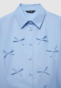 Camicia azzurro chiaro con colletto piegato, caratterizzata da diversi fiocchi di tessuto come dettagli e chiusura con bottoni. Texture liscia, design semplice.