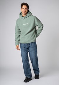 Lichtgroene fleece hoodie met een gestructureerd oppervlak en een wit logo op de borst, gecombineerd met blauwe denim jeans en zwarte sneakers.
