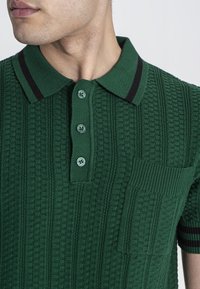 Polo de punto verde con un patrón texturizado, tres botones, un bolsillo en el pecho y rayas negras contrastantes en el cuello.