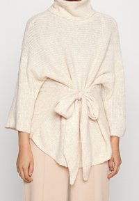 Pull en maille col roulé crème, à manches longues et détail noué à la taille. Tissu texturé avec un motif à tissage ouvert.