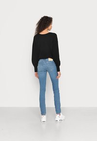 Pulôver oversized preto combinado com jeans skinny azul claro e ténis brancos. Cabelos encaracolados visíveis por trás, com uma etiqueta de logotipo nos jeans.
