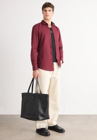 Bordeauxrotes Langarmshirt, schwarze Unterlage, cremefarbene weite Hose, schwarze Ledertasche mit eingeprägtem "HUGO" und schwarze Schuhe. Glatte Texturen überall.