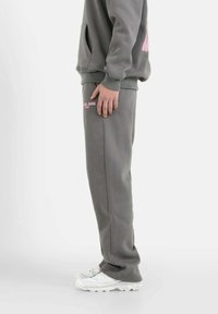 Pantalones de chándal grises en una tela suave, con un logo impreso en rosa en el muslo. El corte es relajado, con bolsillos laterales y puños elásticos.