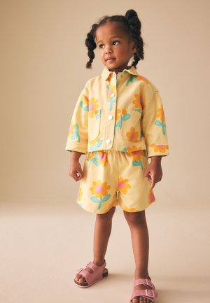 Lille barn med fletninger iført en gul blomsterjakke og shorts sæt, stående med lyserøde sandaler mod en ensfarvet baggrund.