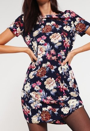 Frau trägt ein marineblaues kurzärmeliges Kleid mit großem Blumenmuster und posiert mit den Händen in der Hüfte.