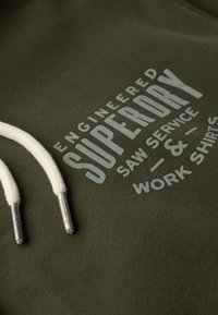 Olijfgroene hoodie met witte trekkoorden en metalen puntjes. Bevat een tekst aan de voorkant in lichtgrijs, met de tekst "Engineered Superdry Saw Service & Work Shirts."