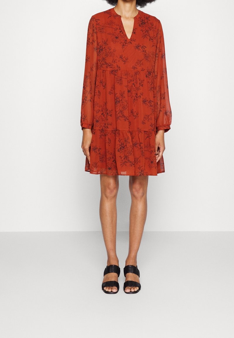 Femme portant une robe rouge mi-longue à manches longues avec un imprimé floral noir et des sandales noires à bouts ouverts, debout devant un fond uni.