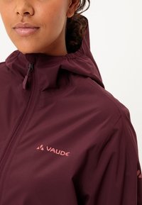 Giacca impermeabile color borgogna con colletto alto, cappuccio regolabile e dettaglio logo rosa. Tessuto liscio e zip frontale.