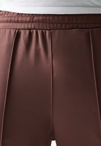 Καφέ αθλητικό παντελόνι με πιέτες, κατασκευασμένο από λείο ύφασμα. Λαστιχένια μέση με ραφές, χωρίς σχέδια ή εμφανή μεταλλικά στοιχεία.