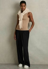 Mujer de pie con las manos en los bolsillos de pantalones negros de pierna ancha, lleva una camiseta sin mangas beige, un suéter crema sobre los hombros y zapatillas blancas.