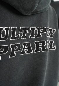 Černá mikina s kapucí, na které je vyšívaný nápis "MULTIPLY APPAREL" v kontrastním bílém stehu. Měkká látka s hladkou texturou a nastavitelnou kapucí.