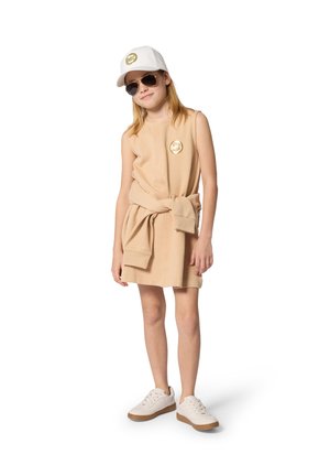 Fille en robe beige sans manches avec un sweat assorti noué autour de la taille, casquette blanche, lunettes aviateur et baskets blanches sur fond blanc.