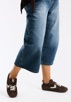 Person wearing cropped blue jeans and brown sneakers with white laces, stepping forward on a white background.