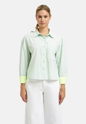 Frau trägt ein hellgrün-weiß gestreiftes Hemd mit neongrünen Manschetten und weiße Hosen mit weitem Bein, steht vor einem schlichten Hintergrund.