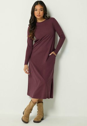 Femme aux longs cheveux foncés portant une robe midi prune à manches longues et des bottines en suède beige, debout devant un fond clair uni.