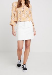 Kvinde iført en gul blomstretdesignet bluse, hvid denim nederdel med frynset kant og sorte sneakers, der går mod en ensfarvet baggrund.