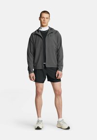 Veste légère grise avec poches zippées, portée sur une chemise noire et un short noir. Le modèle se tient dans une pose neutre avec des chaussures de sport.