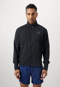Černá sportovní bunda s vysokým límcem, předním zipem a dlouhými rukávy. Obsahuje nenápadné logo a hladký povrch. Krátké modré šortky.