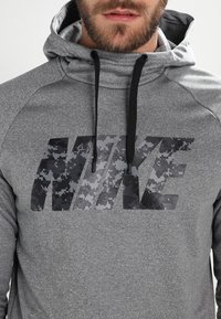 Sweatshirt à capuche gris avec une finition texturée. Présente un grand logo Nike noir et gris en camouflage affiché en évidence sur le devant.