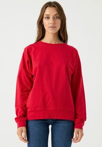 Sudadera roja de cuello redondo con mangas largas, corte relajado y puños de canalé, combinada con jeans ajustados azules. Textura de tela suave.