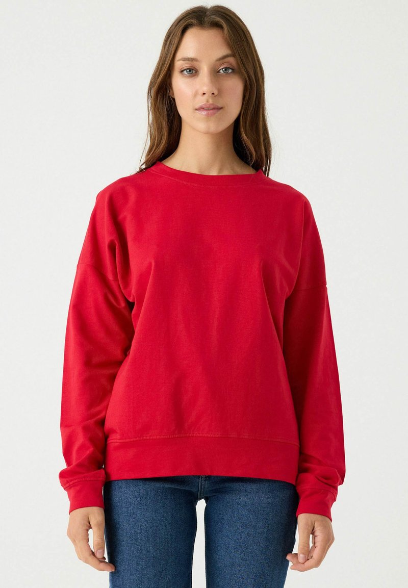 Sudadera roja de cuello redondo con mangas largas, corte relajado y puños de canalé, combinada con jeans ajustados azules. Textura de tela suave.