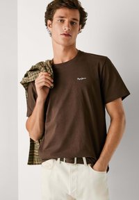 Brun bomulds t-shirt med et lille hvidt logo, parret med cremefarvede bukser og en grøn- og brunternet skjorte hængende over skulderen.